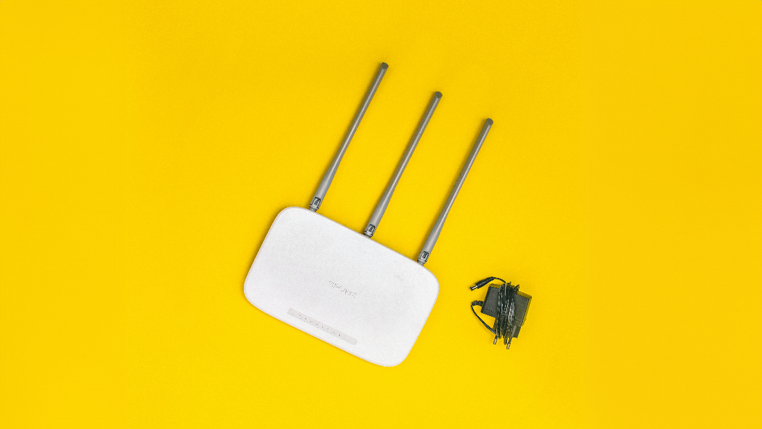 A white WiFi range extender on a yellow background. The range extender has three antennas, a square, white body, and is pictured next to a power cord.