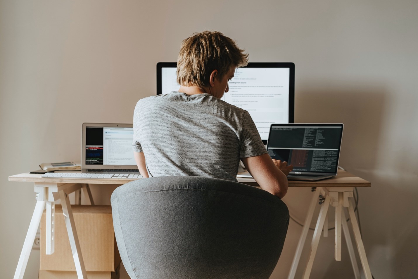 A young male professional works from a minimalist home office setup. On the desk, there are two laptops and one desktop computer. The young man appears to be coding on one of the laptops.